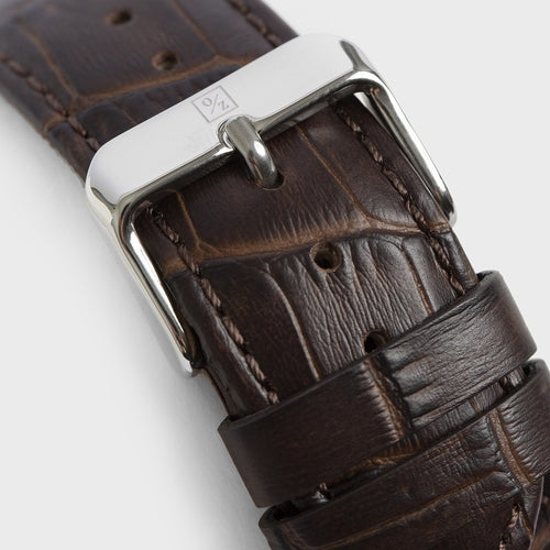 Close-up of a brown leather watch strap with a silver buckle on a light gray background